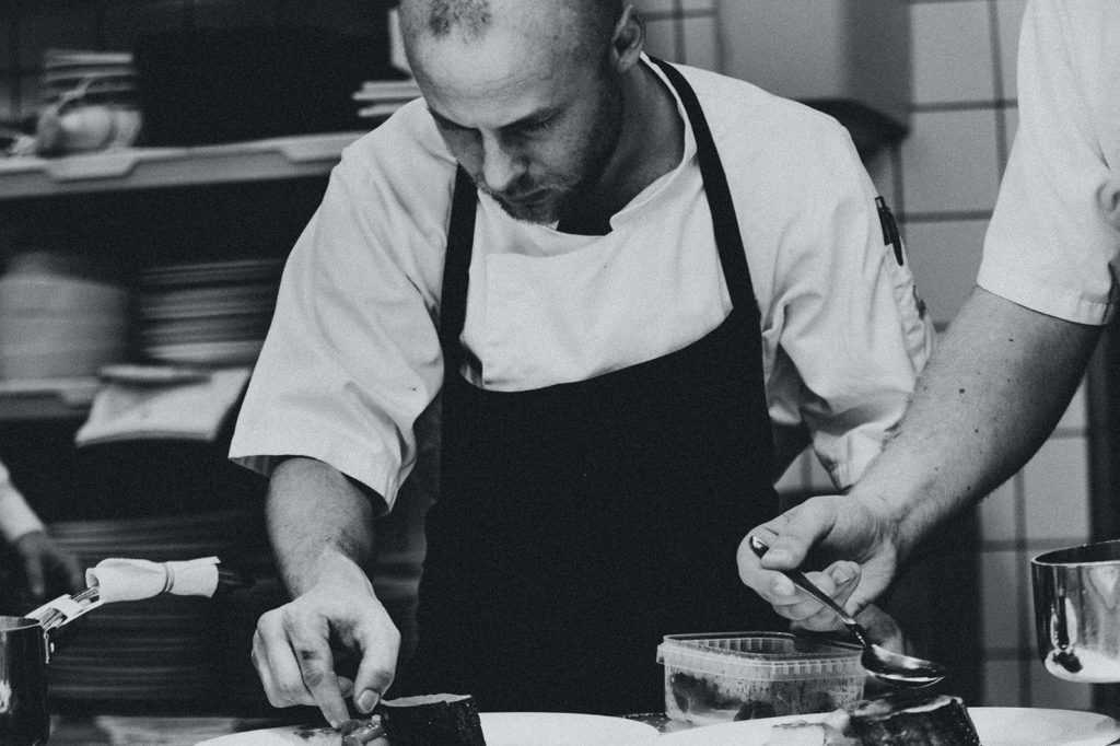 A black and white photo of a chef plating a dish in the kitchen of a restaurant.