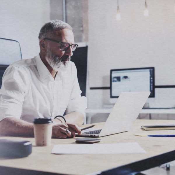 A man with a white and grey beard, wearing glasses and sitting at a desk looking at a laptop.