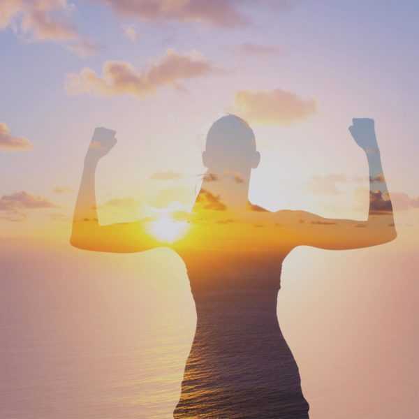 A silhouette of a women flexing both of her biceps with an ocean sunset in the background.